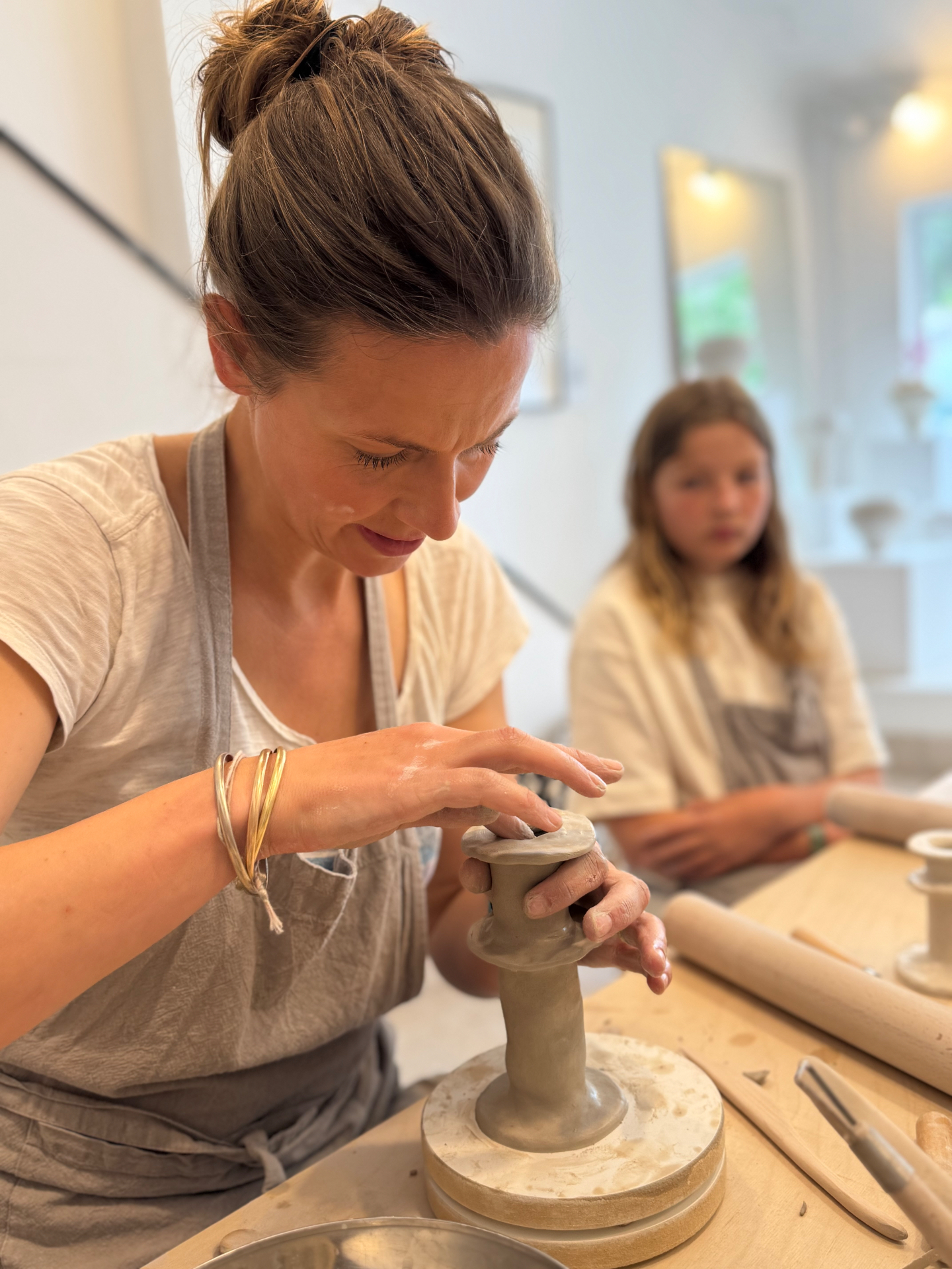A woman shaping a small clay vessel on a pottery wheel, a child watching from behind
