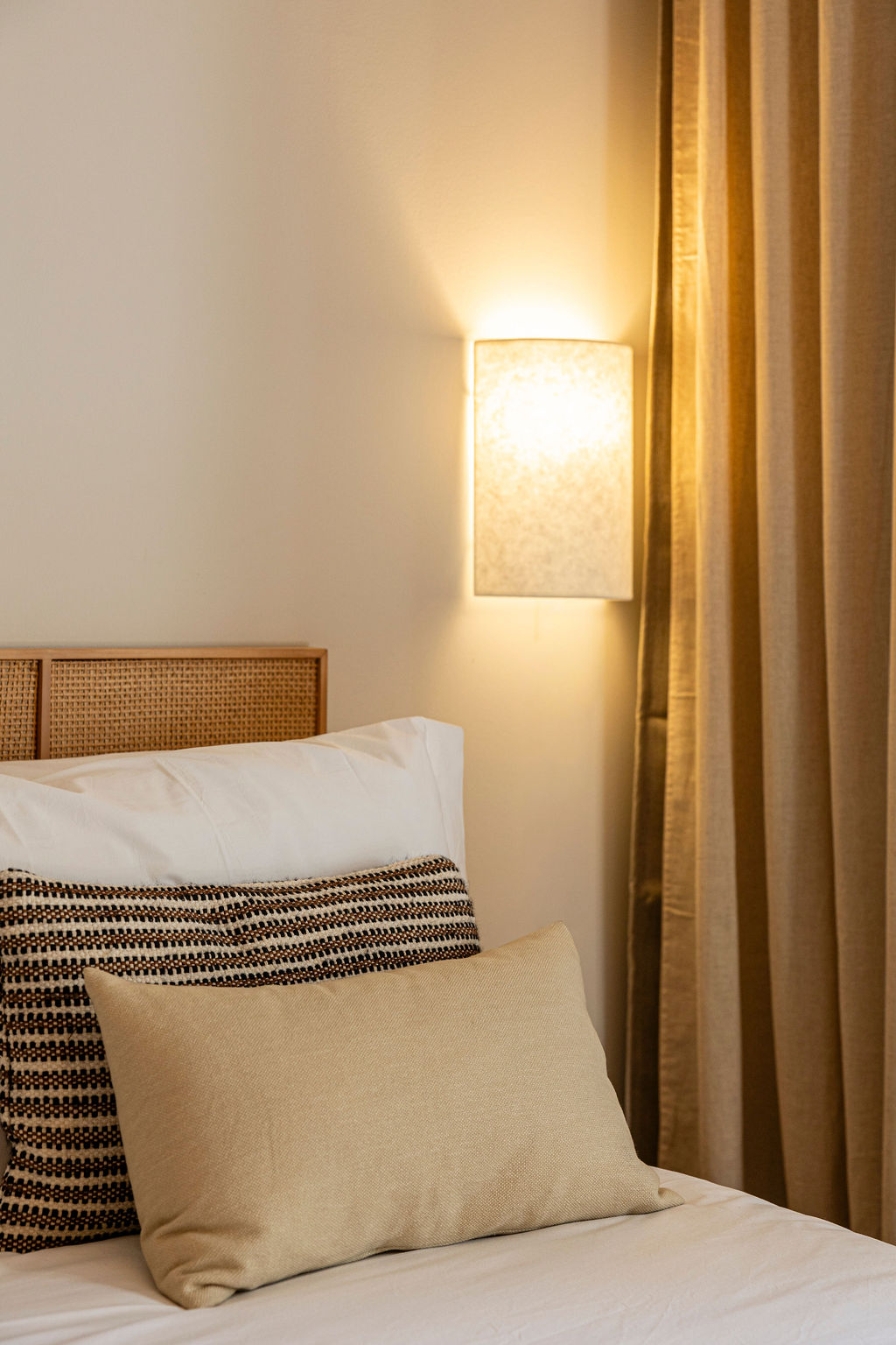 Bedroom one — rattan headboard, linen and a warm wall sconce beside the curtains