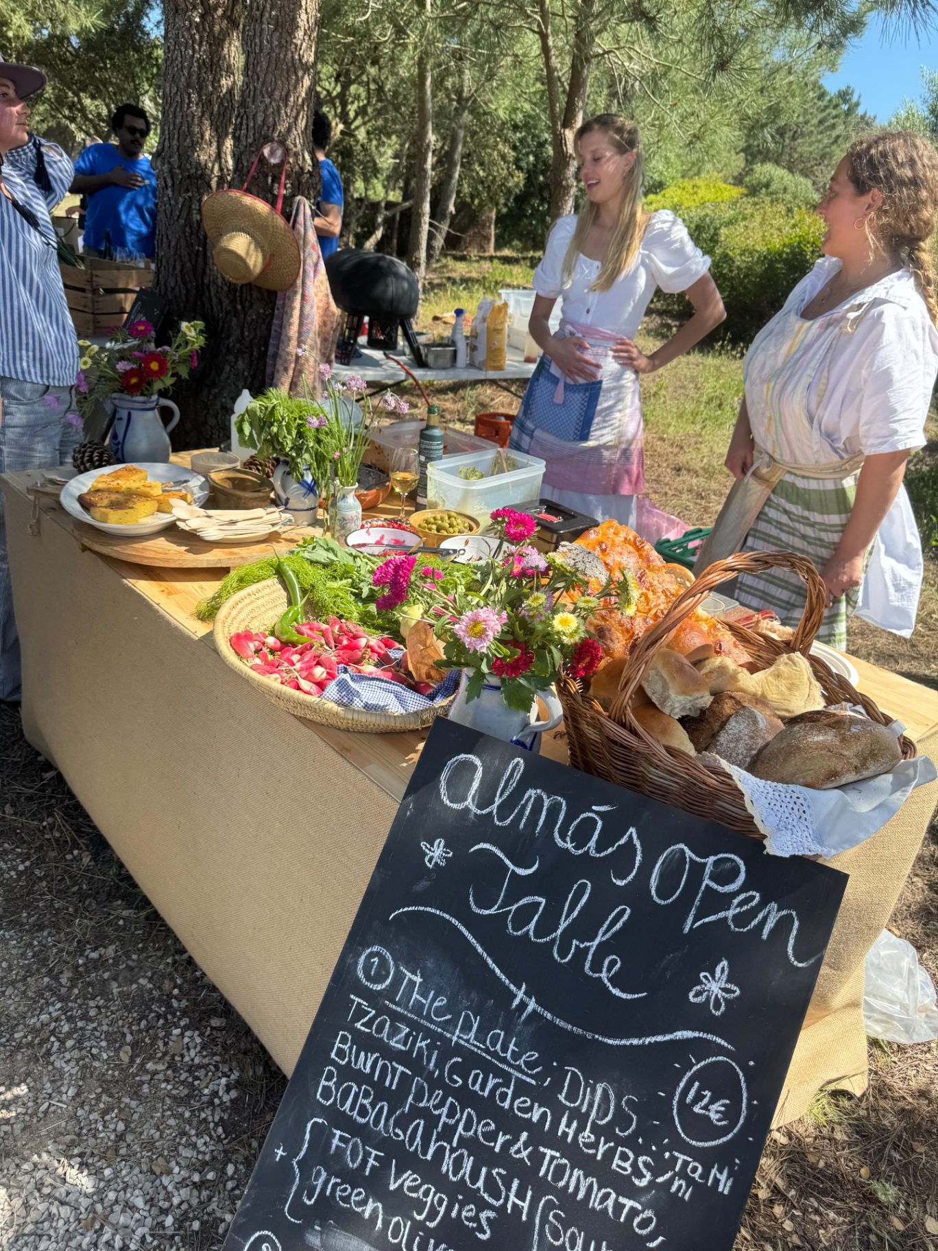 A long open table under the pines set with bread, radishes, salads and wildflowers